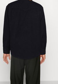 Chemise manches longues bleu marine foncé avec un tissu subtilement texturé, dotée de poignets boutonnés et d'un ourlet arrondi. Portée avec un pantalon vert olive.