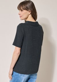 Svart kortärmad skjorta med en texturerad geometrisk mönstring. Kragdesign med rak fåll, matchad med ljusblå jeans.