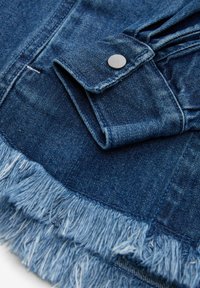 Tessuto in denim blu scuro con bordi sfrangiati, chiusura con bottone e cuciture dettagliate. La texture è robusta con una finitura morbida.