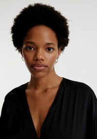 Schwarzes V-Ausschnitt-Oberteil aus weichem Stoff. Das Modell hat kurze, lockige Haare und kleine Creolen, vor einem neutralen Hintergrund.