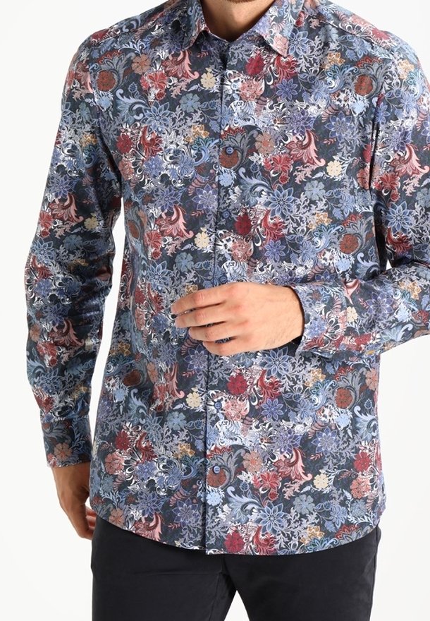 Homme portant une chemise à manches longues boutonnée avec un motif floral multicolore aux tons bleus, rouges et beiges, associée à un pantalon noir.