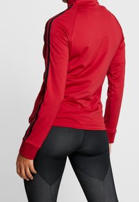Top deportivo rojo con cuello alto, franjas laterales negras y mangas ajustadas, combinado con leggings negros texturizados. Acabado de tela suave.