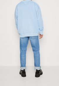 Svetle modrý oversized svetr s žebrovanými manžetami a lemem, v kombinaci se světlomodrými džíny. Modelka má na sobě černé robustní boty.