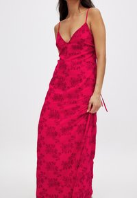 Robe longue rose avec décolleté plongeant, fines bretelles et motifs brodés floraux dans des tons plus foncés, présentant un détail de nœud sur le côté.