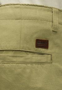 Gros plan sur une poche de pantalon en tissu beige avec un patch logo Timberland marron cousu au-dessus de l'ouverture de la poche.