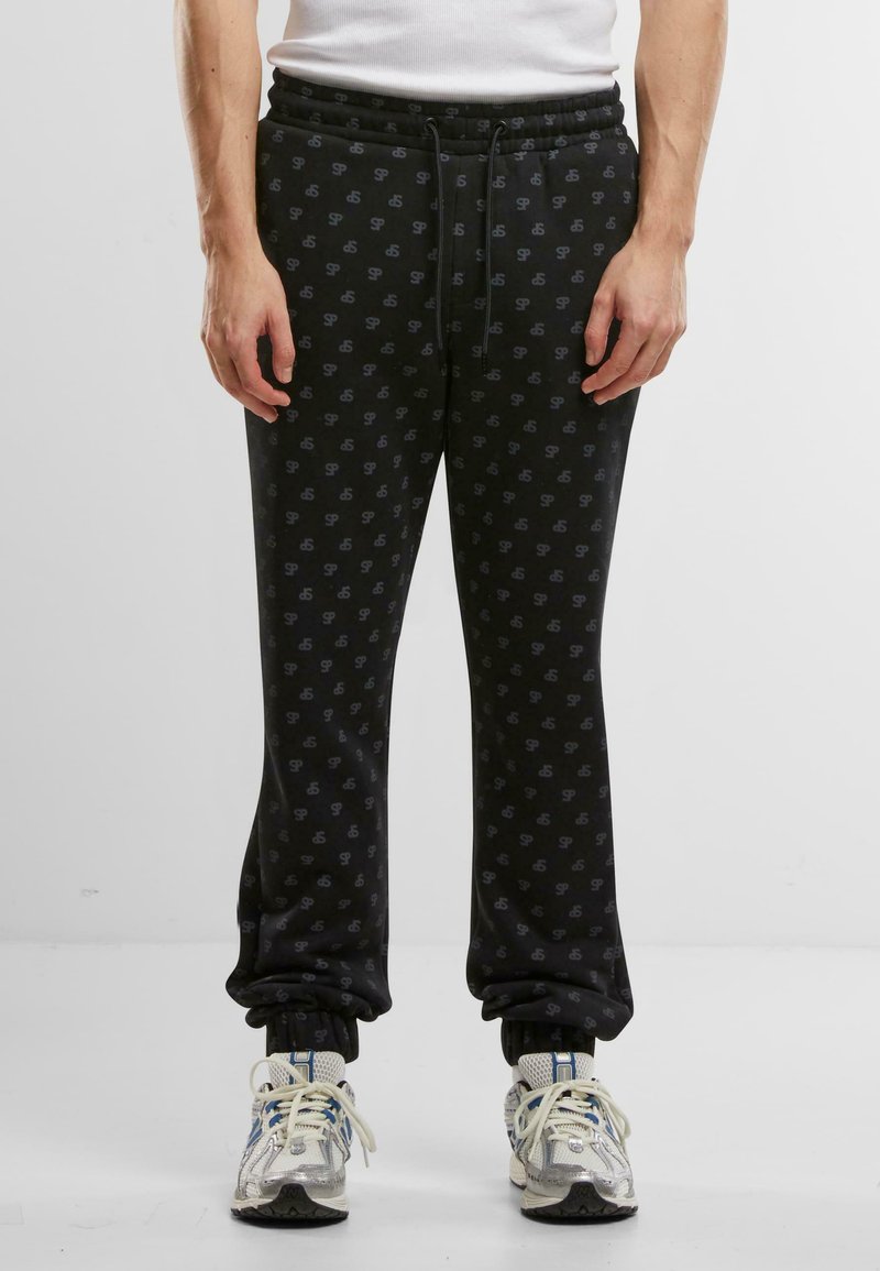 Sorte joggebukser med et subtilt logomønster, elastisk midje og mansjetter. Stoffet ser mykt og behagelig ut. Kombinert med lyse joggesko.