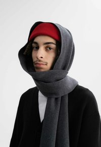 Grijze wollen sjaal om de nek en het hoofd gewikkeld, met een rode gebreide muts. Zwarte cardigan zichtbaar, over een wit shirt gelaagd.