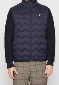 Marinblå pufferväst med vågmönster, hög krage och dragkedja. Har texturerade mörka ärmar och en liten logotyp på bröstet.