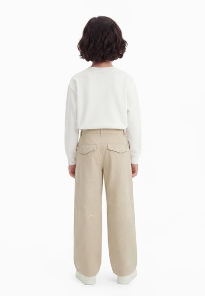 Enfant aux cheveux bouclés et noirs, debout de dos, portant une chemise blanche à manches longues, un pantalon beige à jambes larges avec des éclaboussures de peinture blanche, et des chaussures blanches.