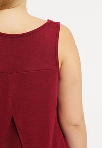 Burgundy ärmlös topp med rund halsringning och en slitsdetalj i ryggen. Den texturerade, ribbade tyget ger en avslappnad passform.