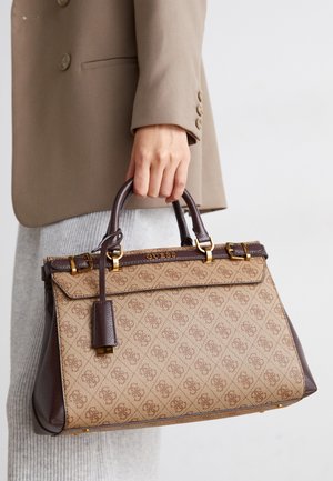 Main tenant un sac à main Guess beige et marron avec motif monogramme, accessoires dorés et poignées foncées, sur un fond neutre.