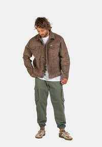 Chaqueta marrón texturizada con dos bolsillos en el pecho, usada sobre una camisa blanca. Combinada con pantalones cargo verdes y zapatillas marrones.