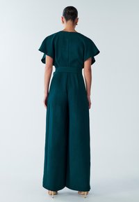 Teal jumpsuit with wide legs, a fitted waist, and flutter sleeves. Smooth fabric with a subtle matte finish and concealed back zipper.
