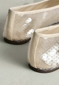 Bēšas baleta kurpes ar caurspīdīgu fliteru pārklājumu, noapaļotu degunu, mīkstu audumu un brūnu gumijas zolīti. Akcenta šuves gar malām.