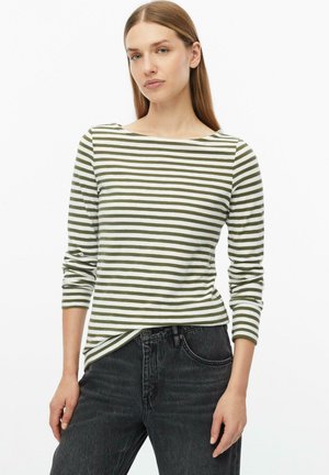 Frau mit glatten Haaren, die ein weiß-olivgrün gestreiftes Langarmshirt und dunkelgraue Jeans trägt, steht mit einer Hand an der Seite.