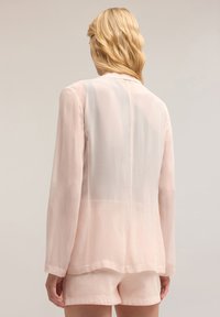 Blazer rosa chiaro con retro trasparente, maniche lunghe e colletto a punte. Texture liscia con leggera lucentezza e design minimalista.
