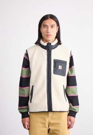 Chaleco de forro polar en color crema, con una cremallera y un bolsillo negro. La camisa de manga larga que lleva debajo presenta rayas verdes y negras, decoradas con rosa.