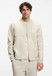 Licht beige bomberjack met geribbelde boorden en ritssluiting. Heeft een gestructureerde stof en een logo op het linker borstgedeelte.