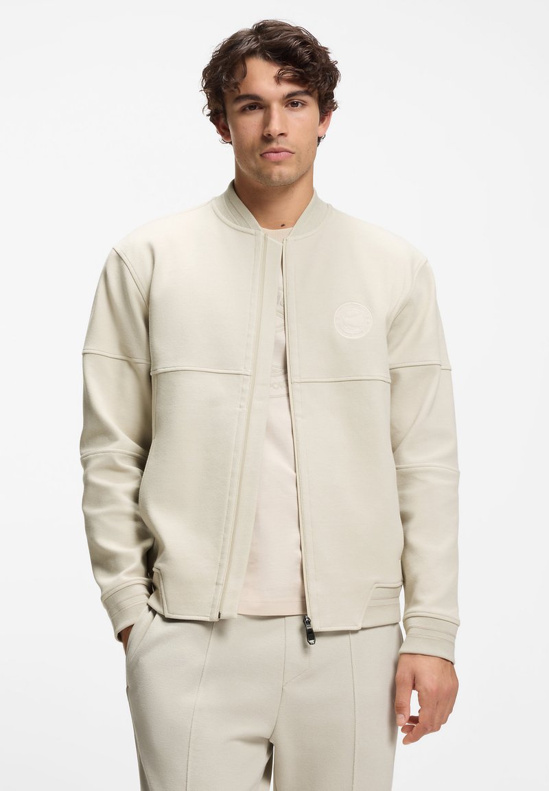 Licht beige bomberjack met geribbelde boorden en ritssluiting. Heeft een gestructureerde stof en een logo op het linker borstgedeelte.