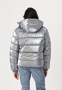 Giubbotto puffo argento con cappuccio, caratterizzato da una texture lucida e liscia, design a righe orizzontali e polsini elastici. Indossato con jeans blu.