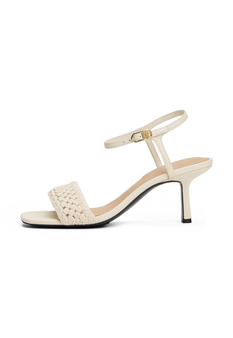 Sandale à talons beige avec une bride tissée et une boucle de cheville réglable. Matériau lisse avec un léger éclat et un talon aiguille élancé.