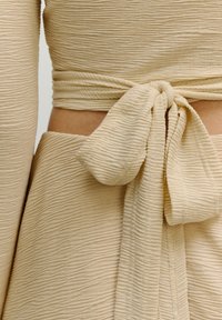 Beiger strukturiert Stoff mit subtilen Wellen, der ein gebundenes Taillendetail aufweist. Der Stoff hat ein weiches, leichtes Aussehen mit einer glatten Oberfläche.