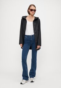 Chaqueta negra impermeable con capucha, llevada sobre una camiseta blanca y jeans acampanados azules. Zapatillas blancas completan el conjunto. Los accesorios incluyen gafas de sol cuadradas.