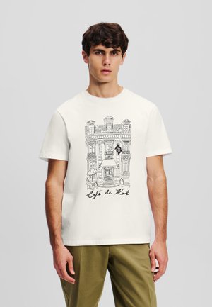 T-shirt in cotone bianco con una linea nera che disegna la facciata di un caffè e la scritta "Café de Karl" sottostante. Design classico a girocollo.
