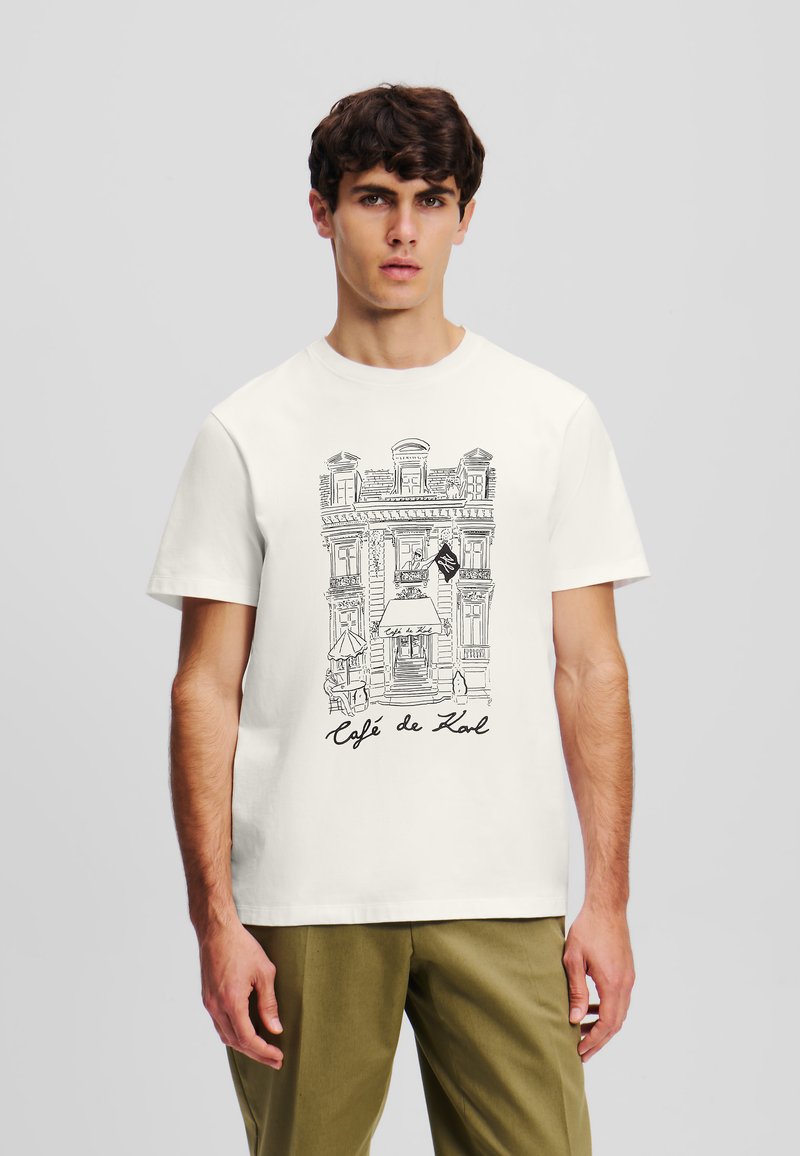Hvid bomulds T-shirt med en sort linjeteckning af en caféfacade med teksten "Café de Karl" under. Klassisk crew neck design.
