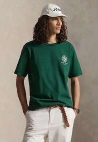 Junger Mann mit lockigem Haar, der ein grünes Ralph Lauren T-Shirt, weiße Hose, beige Ralph-Mütze, geflochtenen braunen Gürtel und Armbanduhr trägt, die Hände in den Taschen.