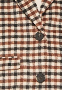 Kariertes Jackett in Creme, Braun und Schwarz. Verfügt über große Knöpfe und einen eingekerbten Kragen. Der Stoff hat eine weiche, strukturierte Oberfläche.