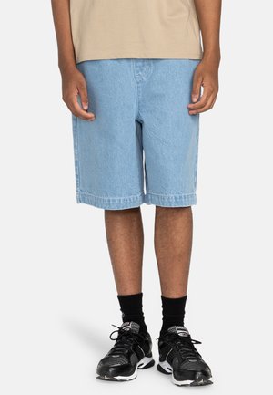 Pantalones cortos de mezclilla azul claro con un ajuste relajado y dobladillo doblado. Combinados con zapatillas deportivas negras y calcetines negros hasta la altura del tobillo.