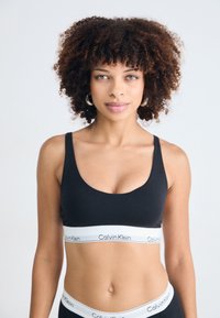 Donna con capelli ricci che indossa un reggiseno sportivo nero di Calvin Klein e una mutandina coordinata, in piedi di fronte a uno sfondo bianco uniforme.