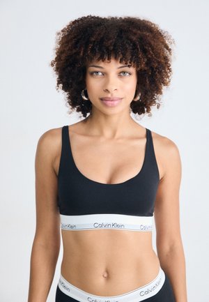 Vrouw met krullend haar, gekleed in een zwarte Calvin Klein sportbh en bijpassend ondergoed, staande tegen een eenvoudige witte achtergrond.