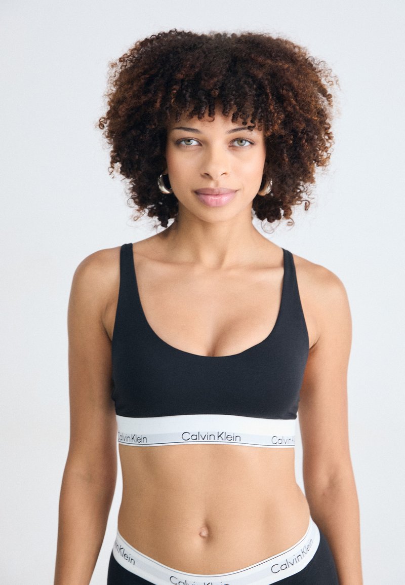 Donna con capelli ricci che indossa un reggiseno sportivo nero di Calvin Klein e una mutandina coordinata, in piedi di fronte a uno sfondo bianco uniforme.