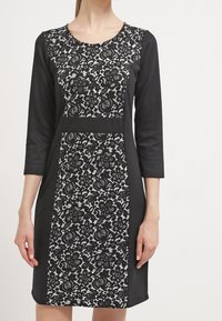 Svartvit klänning med rund halsringning, trekvartslånga ärmar, med spetsmönster i mitten och solida svarta sidor.