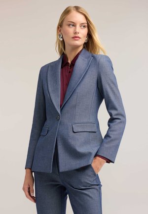 Donna bionda che indossa un blazer blu su misura e pantaloni con una camicia bordeaux con bottoni, in piedi con una mano in tasca, guardando di lato.