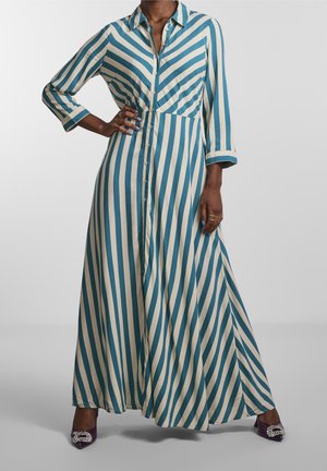 Femme portant une longue robe boutonnée rayée verticalement bleu et blanc, manches trois quarts et talons violets ornés de bijoux.