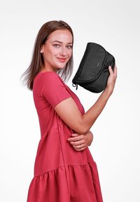 Černá taška vyrobená z látky s jemnou texturou, s zakřiveným designem a zipovým uzávěrem. Model drží tašku proti jednoduchému pozadí.