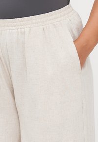 Light beige trousers with a textured, woven fabric, featuring an elastic waistband and side pockets. Relaxed fit with a smooth finish.