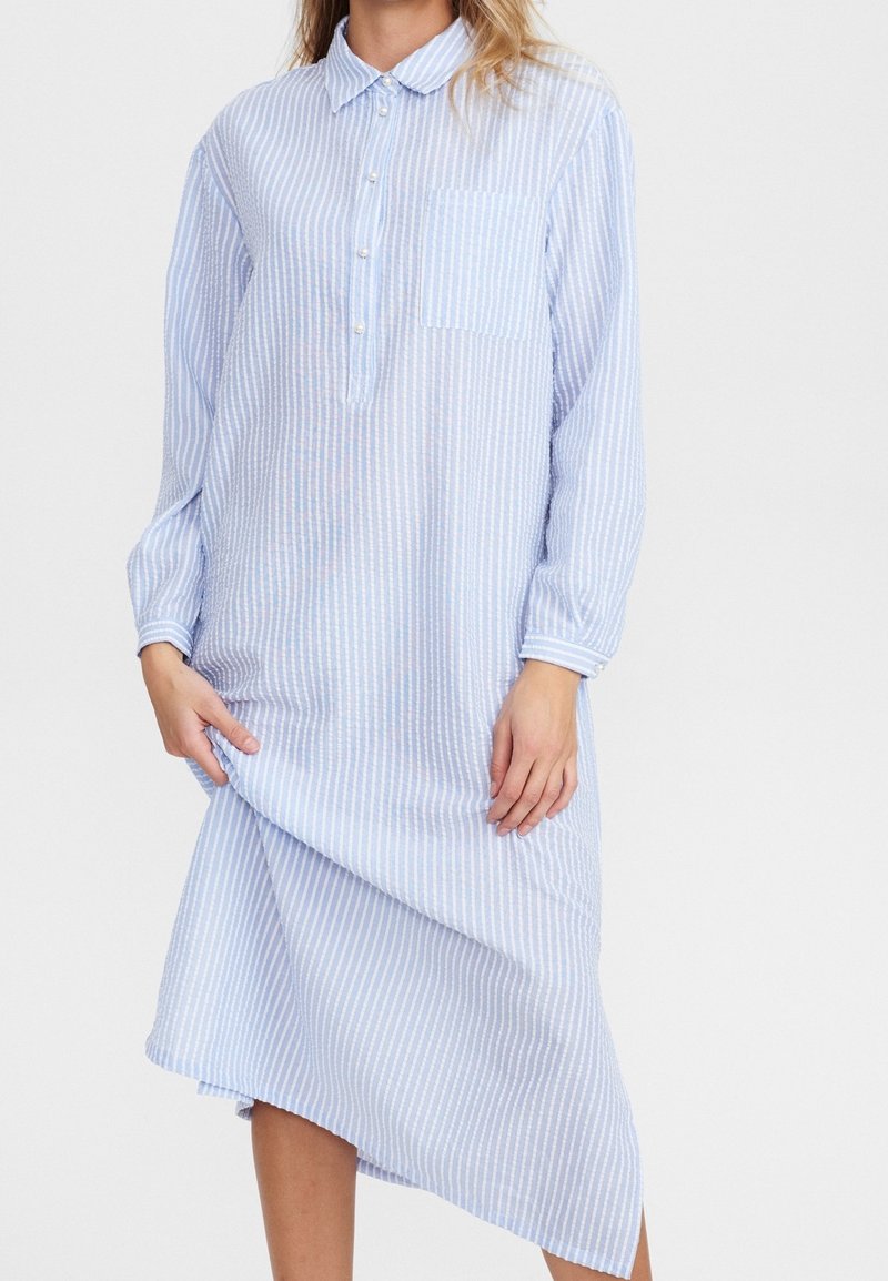 Femme portant une robe chemise à manches longues, rayée bleu clair et blanche, avec une poche avant et un col boutonné, tenant l'ourlet.