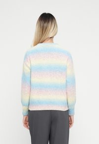 Άτομο με μακριά ombre μαλλιά, φορώντας ένα φλίς παλτό σε παστέλ ριγέ σχέδιο σε μπλε, ροζ και κίτρινο, συνδυασμένο με σκούρες γκρίζες παντελόνες, στραμμένο μακριά.