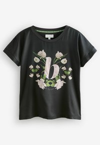 Černé tričko s krátkým rukávem a květinovým vzorem, které má výrazné růžové písmeno "b" obklopené bílými květy a zelenými listy.