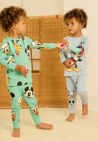 Twee kinderen dragen bijpassende pyjama's met prints van tekenfilmfiguren. De ene outfit is groen met Mickey en Donald, en de andere is lichtblauw met Mickey en Goofy.