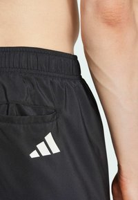 Czarne spodenki sportowe mają elastyczny pas, boczną kieszeń oraz biały trójkątny logo na dolnej lewej stronie. Gładka tekstura materiału.