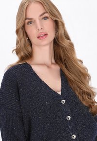 Marine gebreide cardigan met V-hals, versierd met metallic stippen. Bevat vijf zilveren knopen en een textuurdetail op de schouders.