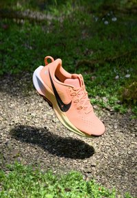 Botte de course rose clair avec un dessus respirant, un swoosh noir de Nike, une semelle texturée et des accents réfléchissants. Flottant au-dessus de gravier et de verdure.