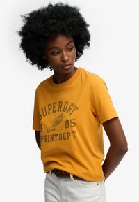 Geel katoenen T-shirt met bedrukt ontwerp met de tekst "Superdry" en een vleugel schoen ontwerp. Korte mouwen en een ronde hals.