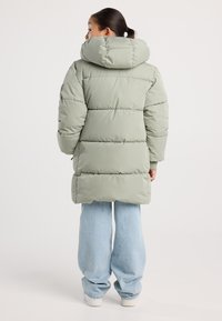 Mujer de espaldas vestida con un abrigo acolchado con capucha en verde salvia, jeans de pierna ancha en azul claro y zapatillas blancas sobre un fondo liso.