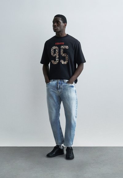 Homme portant un t-shirt noir avec l'inscription léopard "95" et un jean bleu délavé, debout devant un fond gris clair uni.